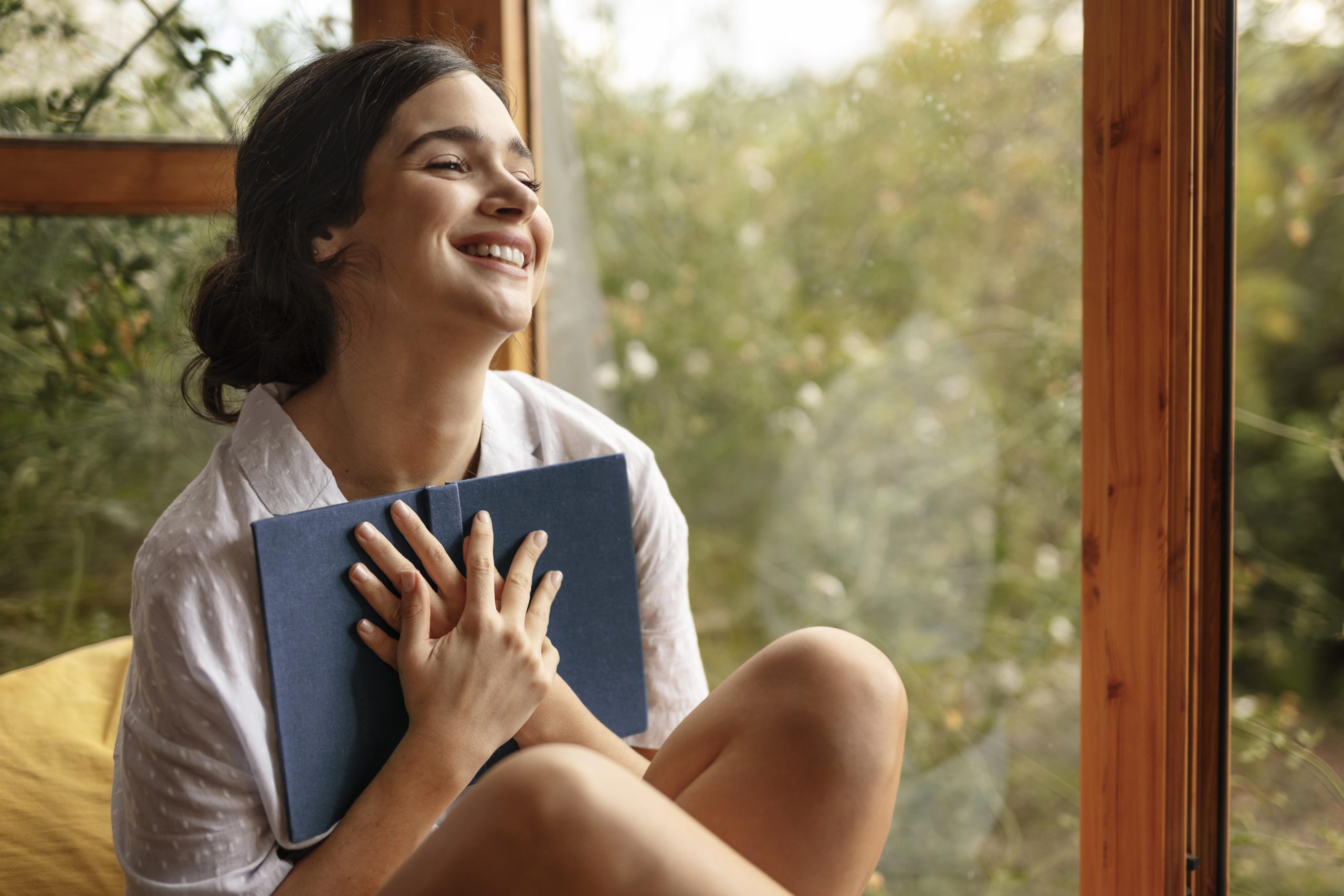 A person looking out of the window and smiling while holding a book against their chest, indicating practising self care tips for mental health.