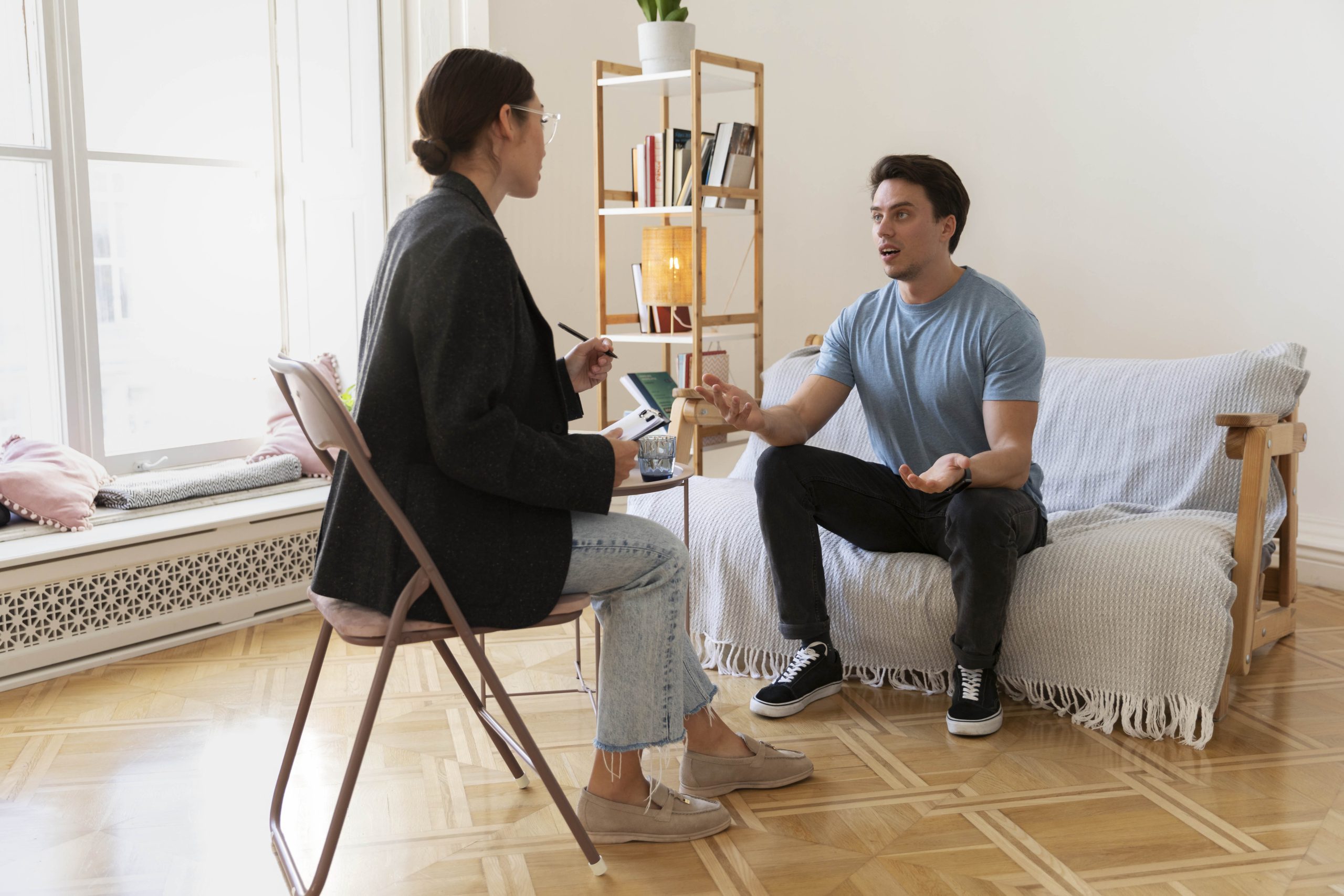 A client sitting on a sofa opposite to the therapist who is sitting in a chair. The client asks questions to determine the right Therapist for their needs.