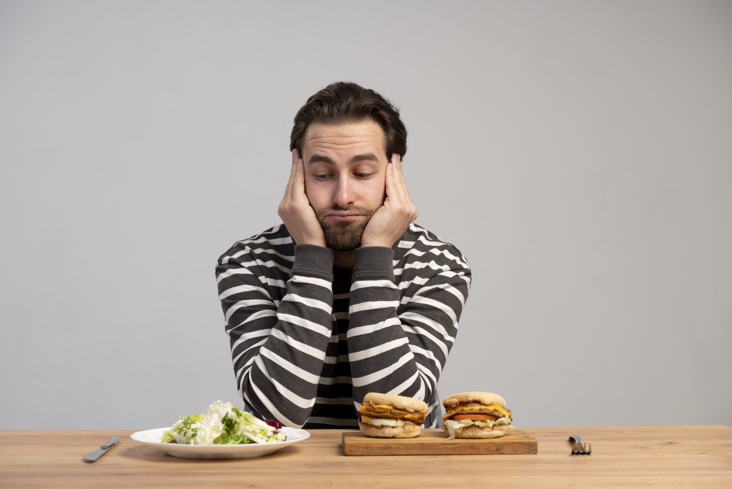 A person sitting at a table, struggling to decide between a plate of salad and a plate of burgers, indicates a common mental health issue.