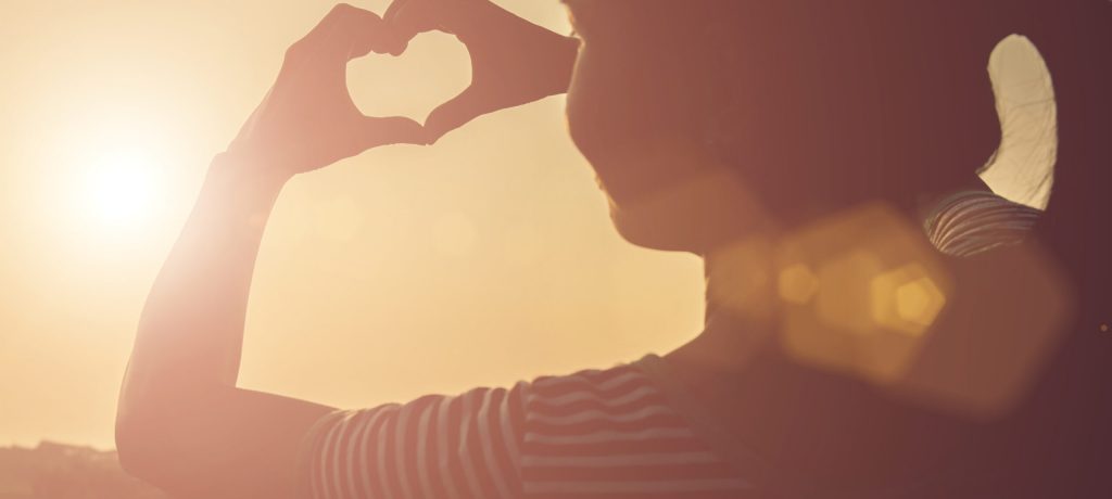 Young woman hands shaped heart against beautiful sunset.